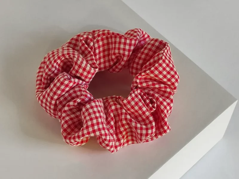 Scrunchie a quadri rosso, realizzato a mano, poggiato su espositore quadrato bianco.