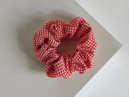 Scrunchie a quadri rosso, realizzato a mano, poggiato su espositore quadrato bianco.