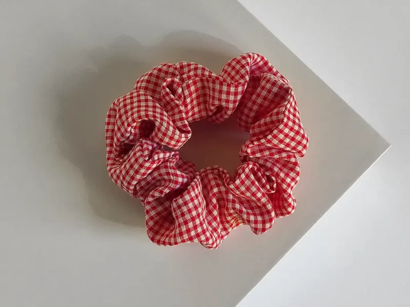 Scrunchie a quadri rosso, realizzato a mano, poggiato su espositore quadrato bianco.